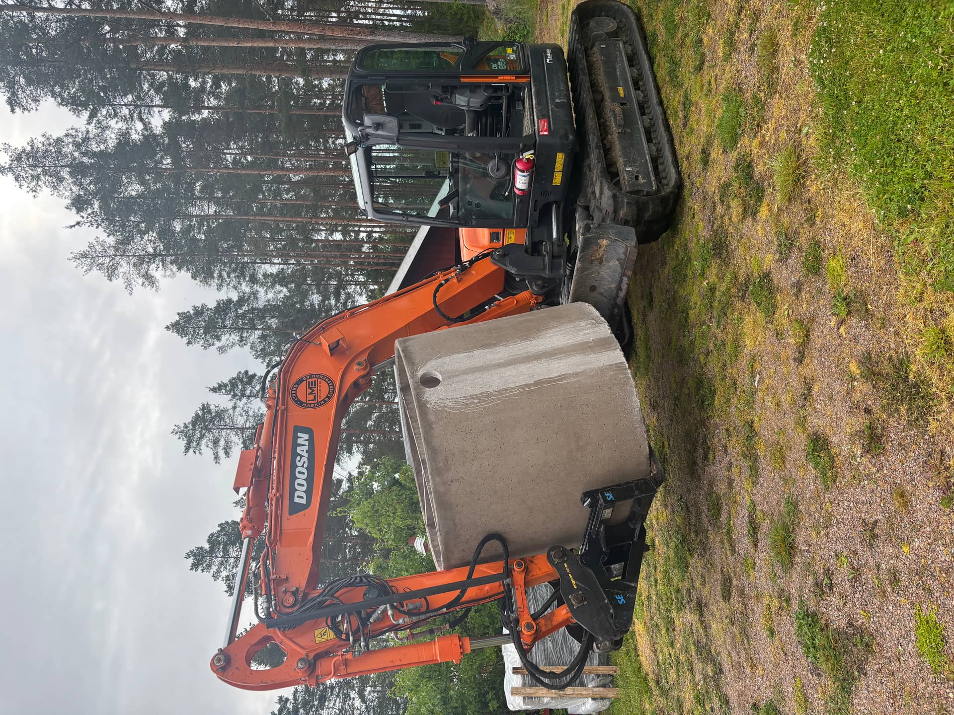 Doosan-grävmaskin som lyfter en grå betongbrunnsring vid tallskog